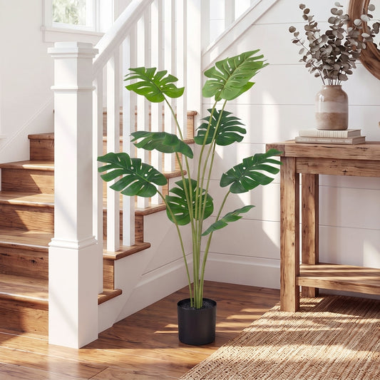 The artificial monstera plant in a black pot placed on a rug next to a wooden table and stairs