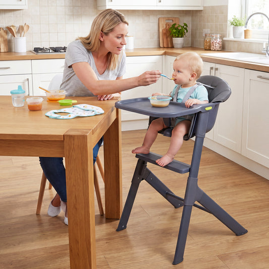 The high chair being used in a kitchen while a mother feeds her baby