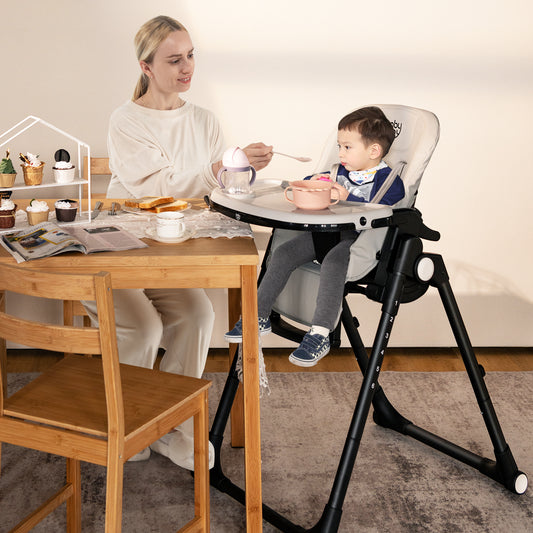 A woman feeding a baby in the grey high chair at a wooden dining table