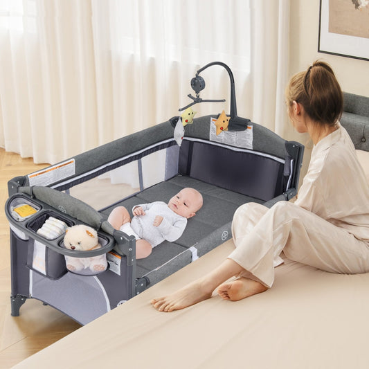 The travel cot with a baby lying inside and a woman sitting beside it