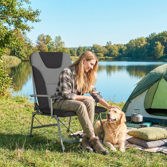 A woman and a dog sit on a padded camping chair next to a tent by a lake