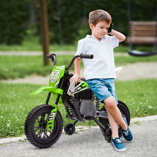 A young boy in a white t-shirt and denim shorts sits on the green kids electric motorbike outdoors