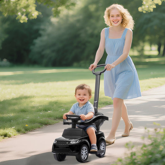 A smiling toddler sitting on the black ride on car being pushed by a woman on a park path