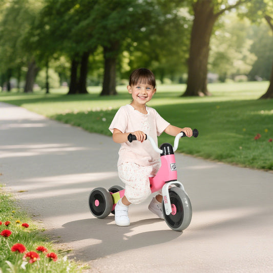 A smiling toddler rides the pink balance bike with no pedals along a paved path in a park
