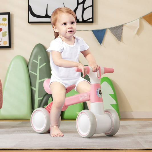 A toddler sits on the pink and white balance bike in a room with decorative trees