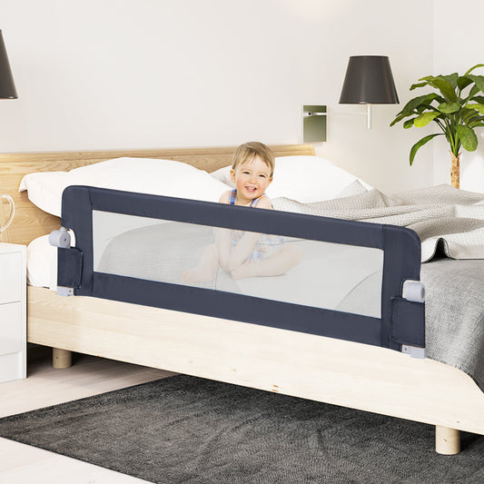 A toddler sits on a bed with the grey bed rail installed, looking towards the camera