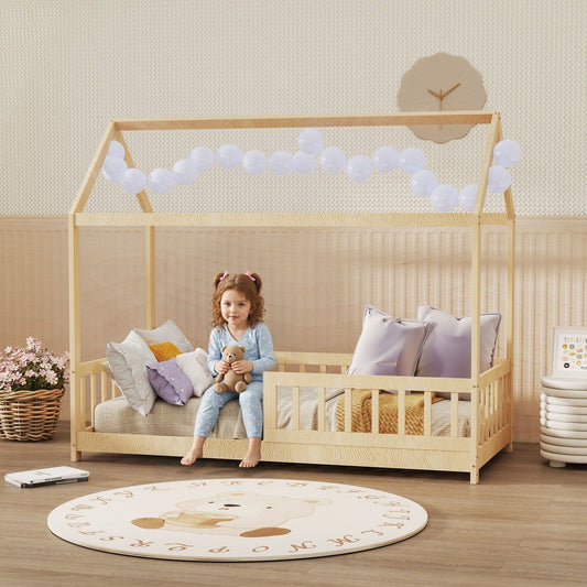 A young girl sits on the natural wood house-shaped kids bed frame in a decorated bedroom