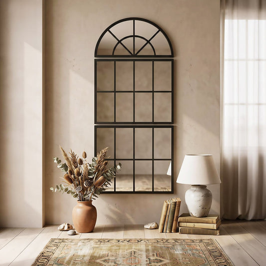 The arched wall mirror with metal grid displayed in a living room setting with a vase of dried flowers and books