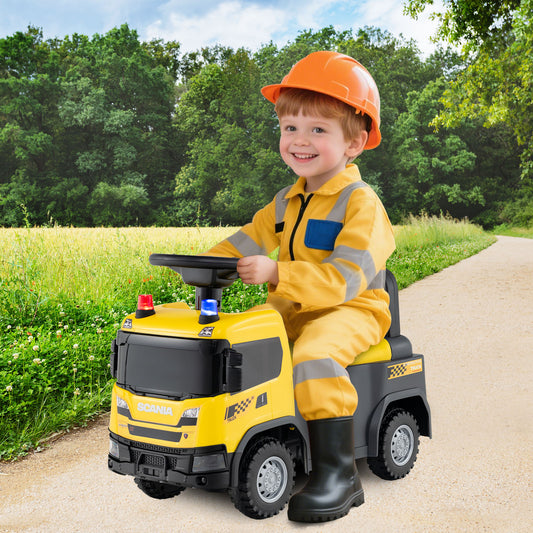A young boy wearing an orange hard hat and yellow overalls rides the yellow push car outdoors on a path