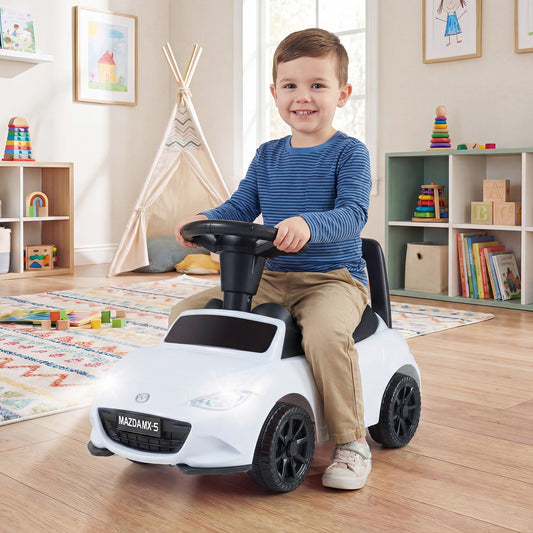 A happy boy rides the white Mazda push car indoors in a playroom