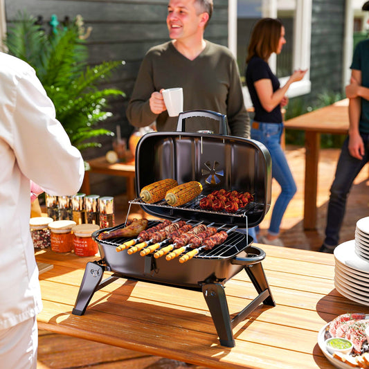 The charcoal grill placed on a wooden table with food cooking while people socialise nearby