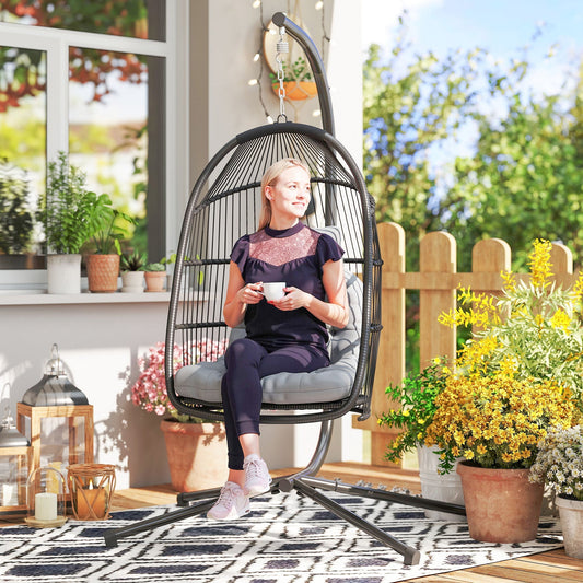 A woman sits in the dark grey rattan hanging egg chair with a cushion on a patio