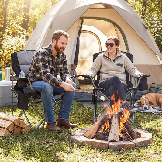 A couple sitting in grey camping chairs by a campfire outside a tent