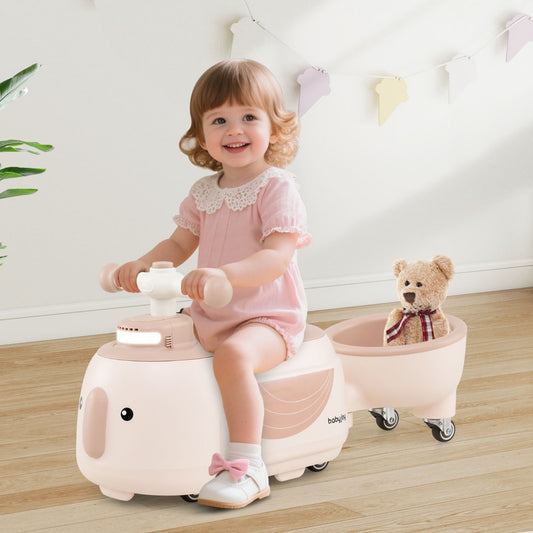 A happy toddler girl sits on the beige ride on toy with trailer, playing with a teddy bear