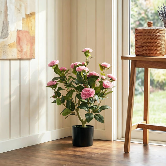 Pink artificial camellia plant in a black pot on a wooden floor next to a table