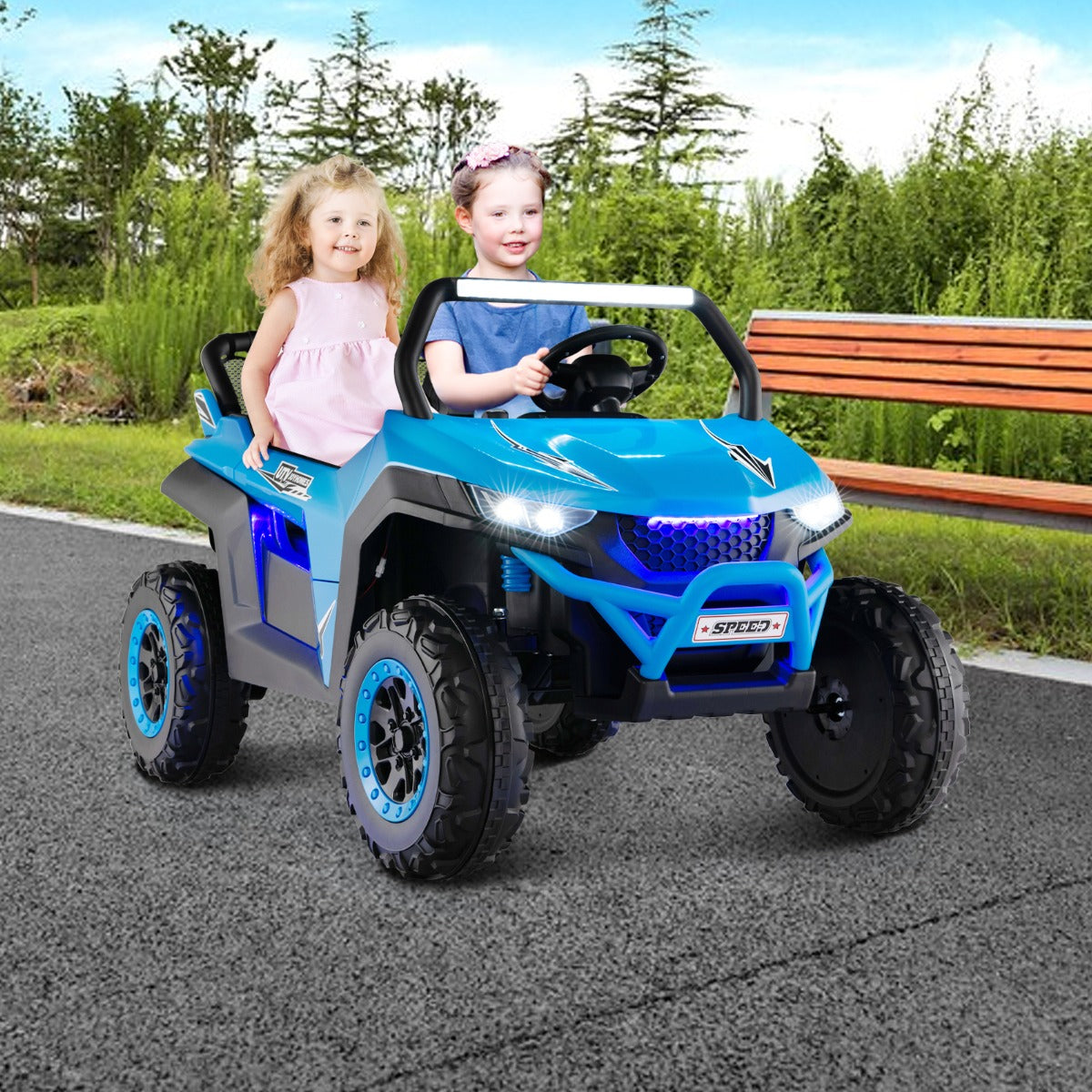 The kids electric car being driven by two children on a paved path near a wooden park bench