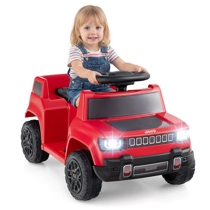 A toddler sits in the red kids electric car, smiling at the camera, parked on grass