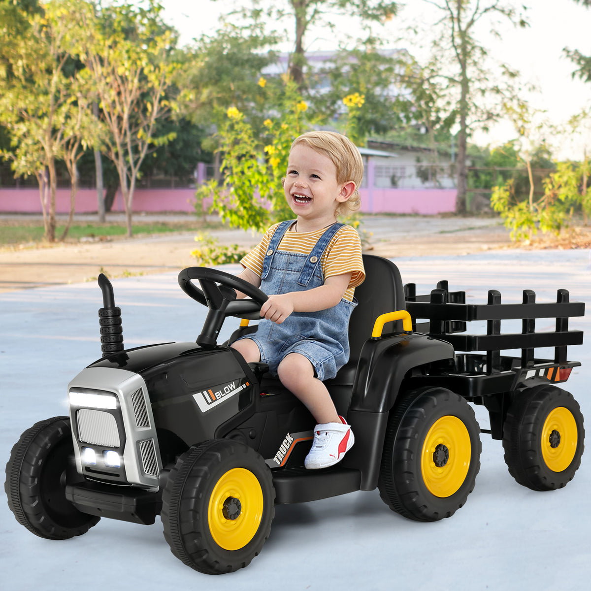 A happy young boy driving the kids electric tractor with a trailer in an outdoor park setting