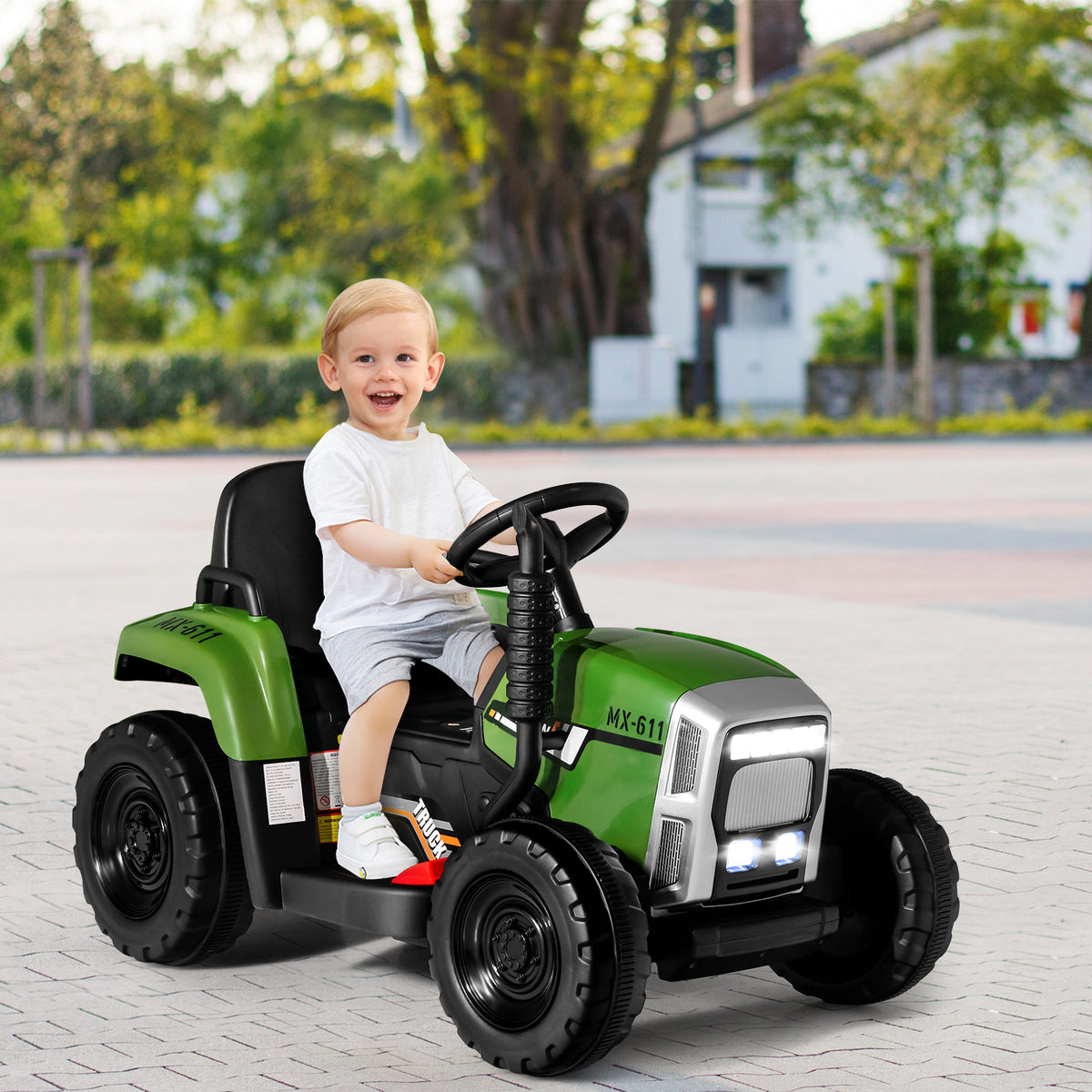 A happy young boy sitting on the kids electric tractor while driving on a paved driveway