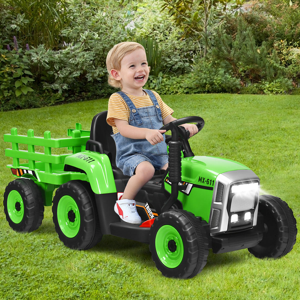 A happy toddler drives the green kids electric tractor with trailer in a garden