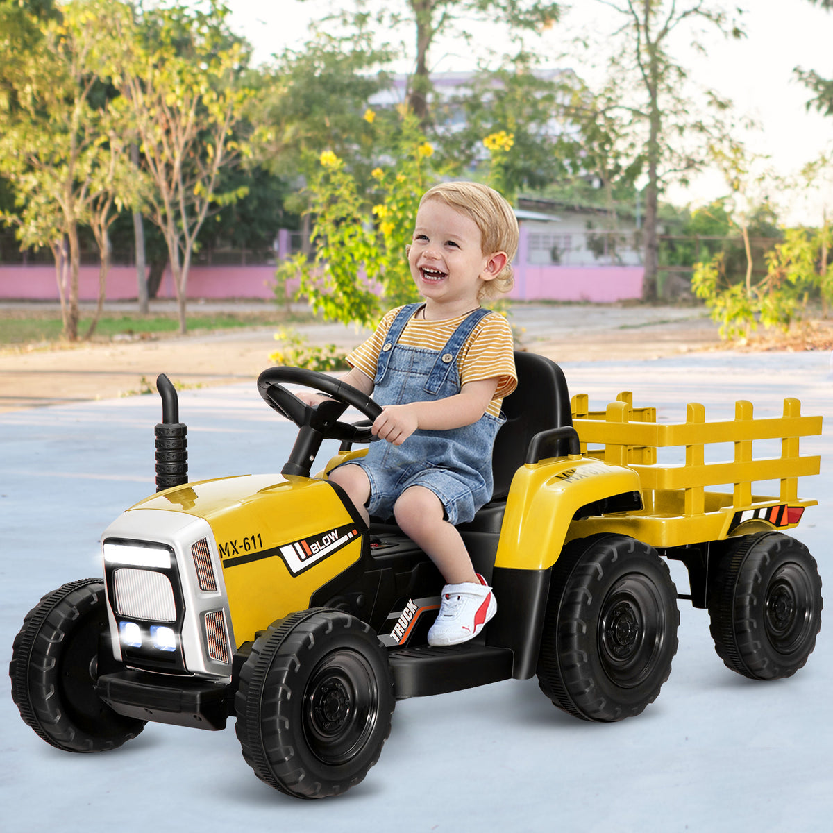 A happy toddler sits in the driver's seat of the yellow ride-on tractor, steering it outdoors
