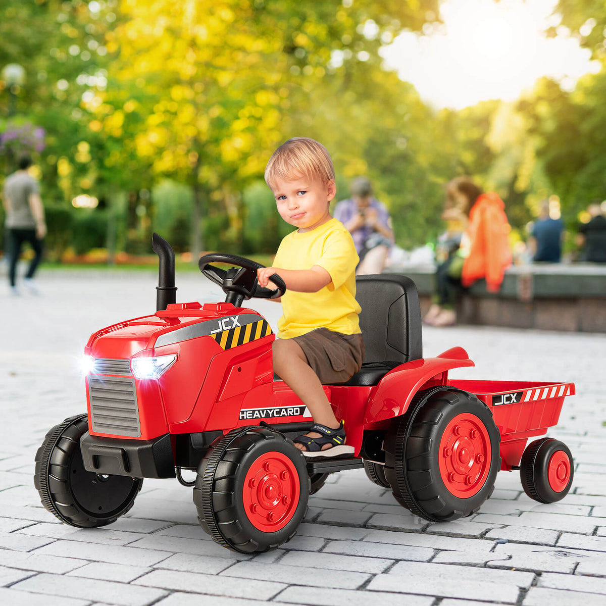 A young boy drives the red ride on tractor with trailer on a paved path