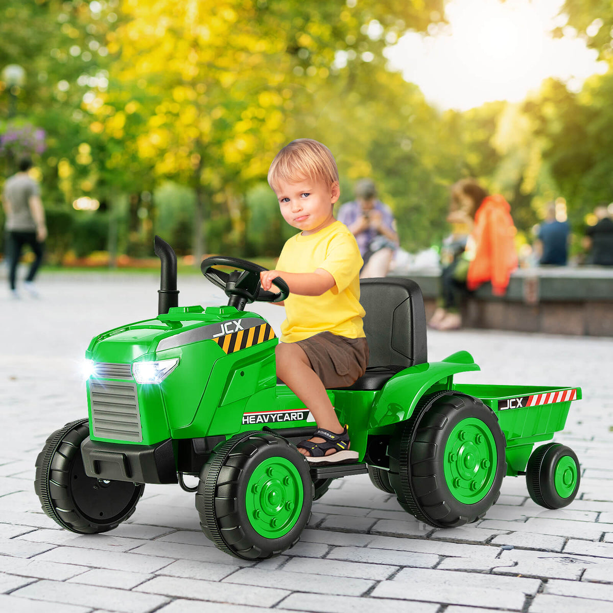 The green kids electric tractor with a trailer being sat on by a young boy on a paved surface