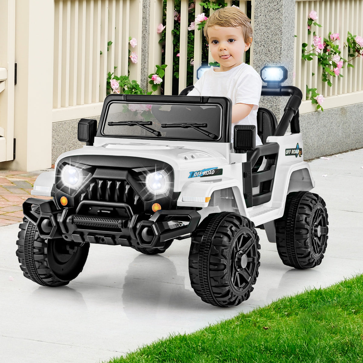 A young boy sits in the white kids electric truck on a paved path with a fence and plants