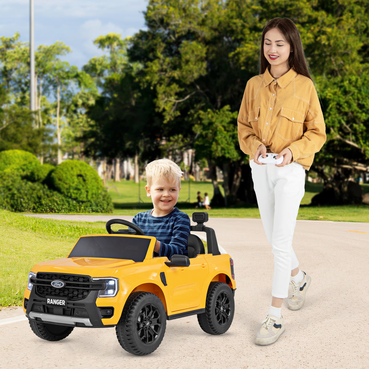 A smiling boy drives the yellow kids electric car while a woman stands nearby holding the remote control