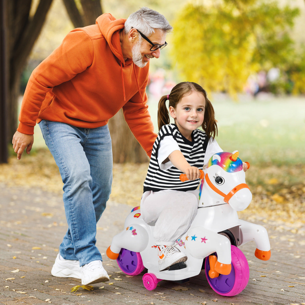 A smiling grandfather pushes a young girl on a white unicorn ride on toy with training wheels in a park