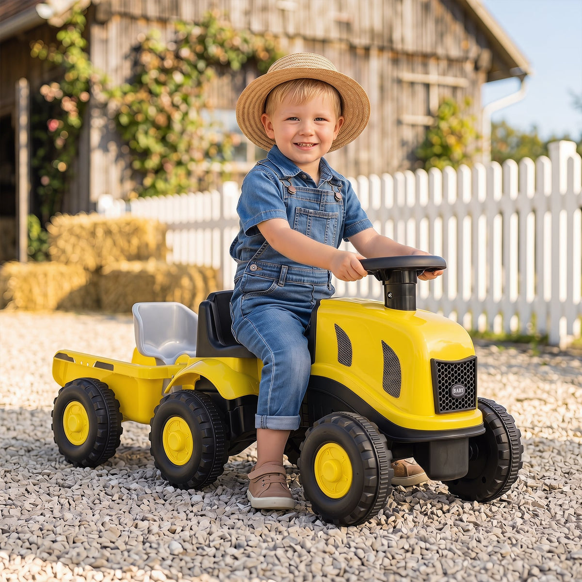 The yellow ride on toy being used by a smiling child in a gravel driveway near a wooden barn
