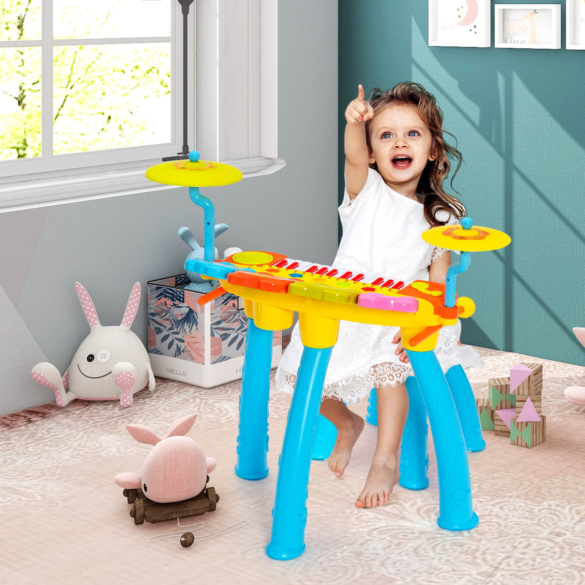 A young girl sits at the blue and yellow toy keyboard drum set, playing with a drumstick