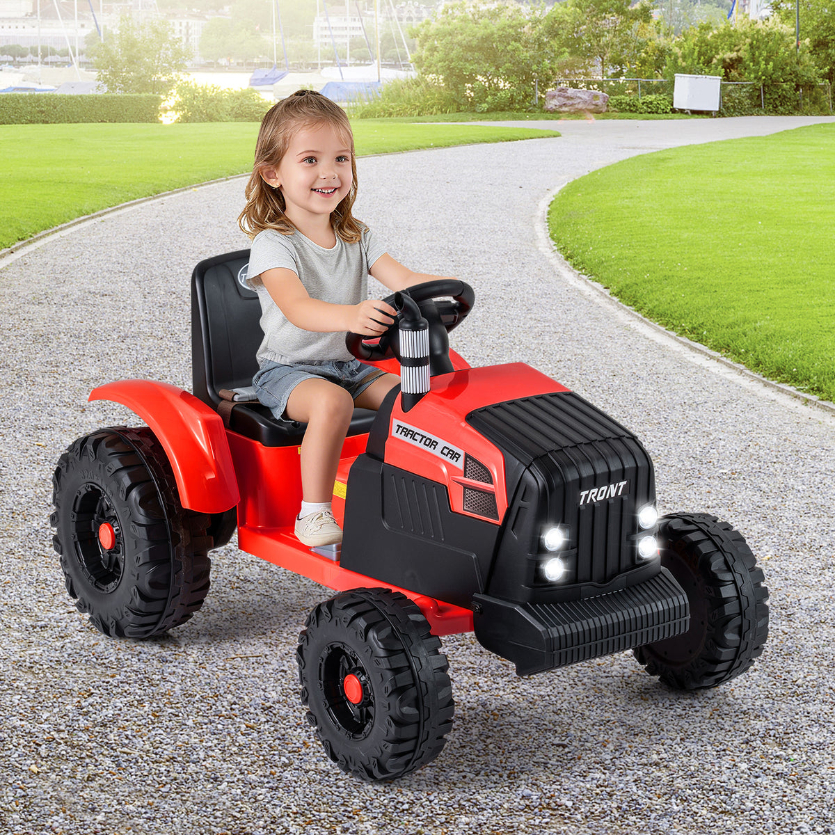 A happy child steers the red ride on tractor with trailer across a paved garden path