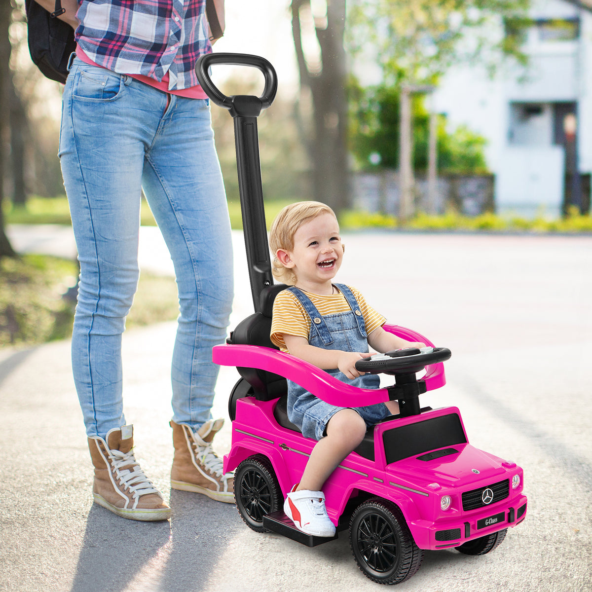 A smiling baby sits in the pink ride-on toy car, pushed by a woman