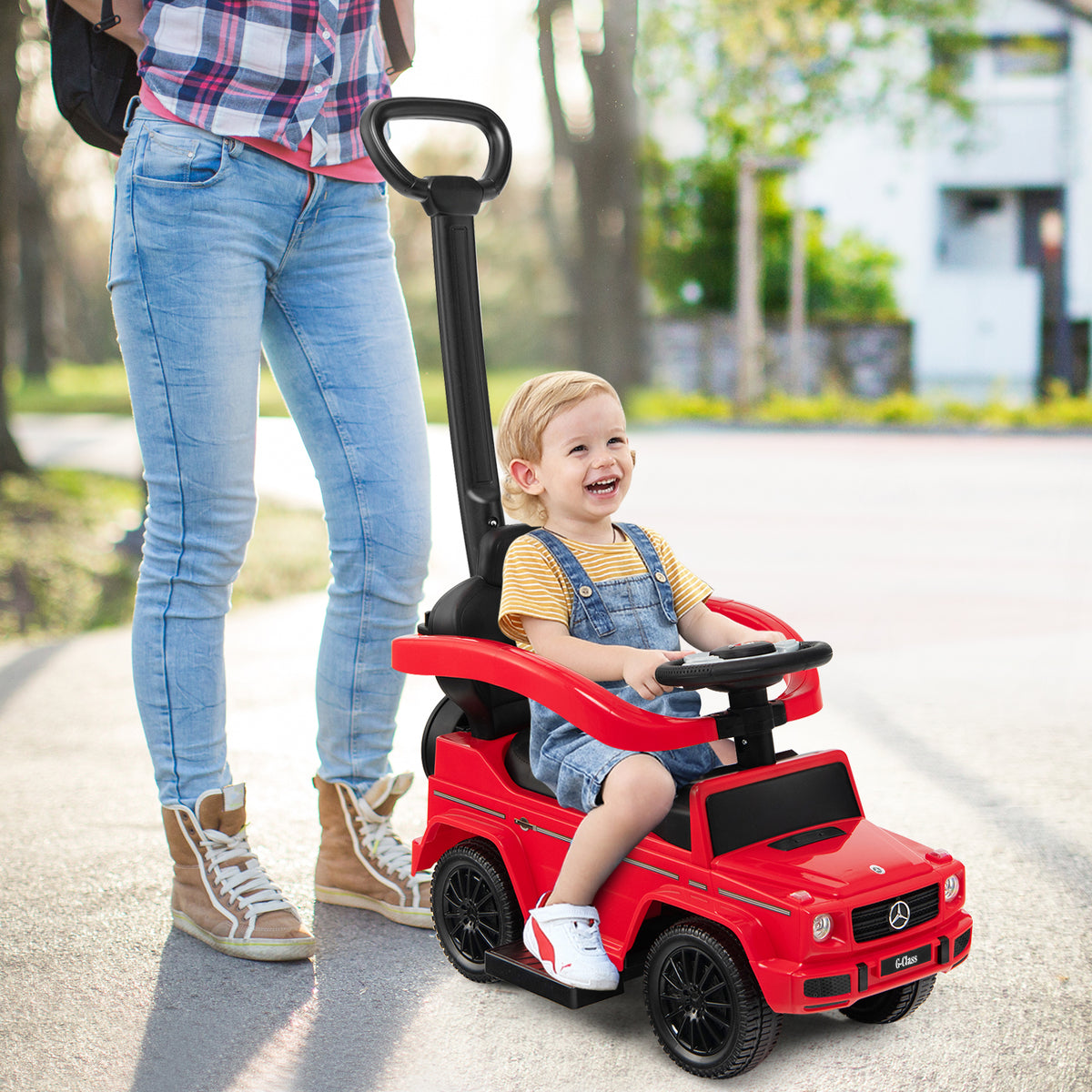 The red baby walker with a push handle being steered by a smiling toddler outdoors