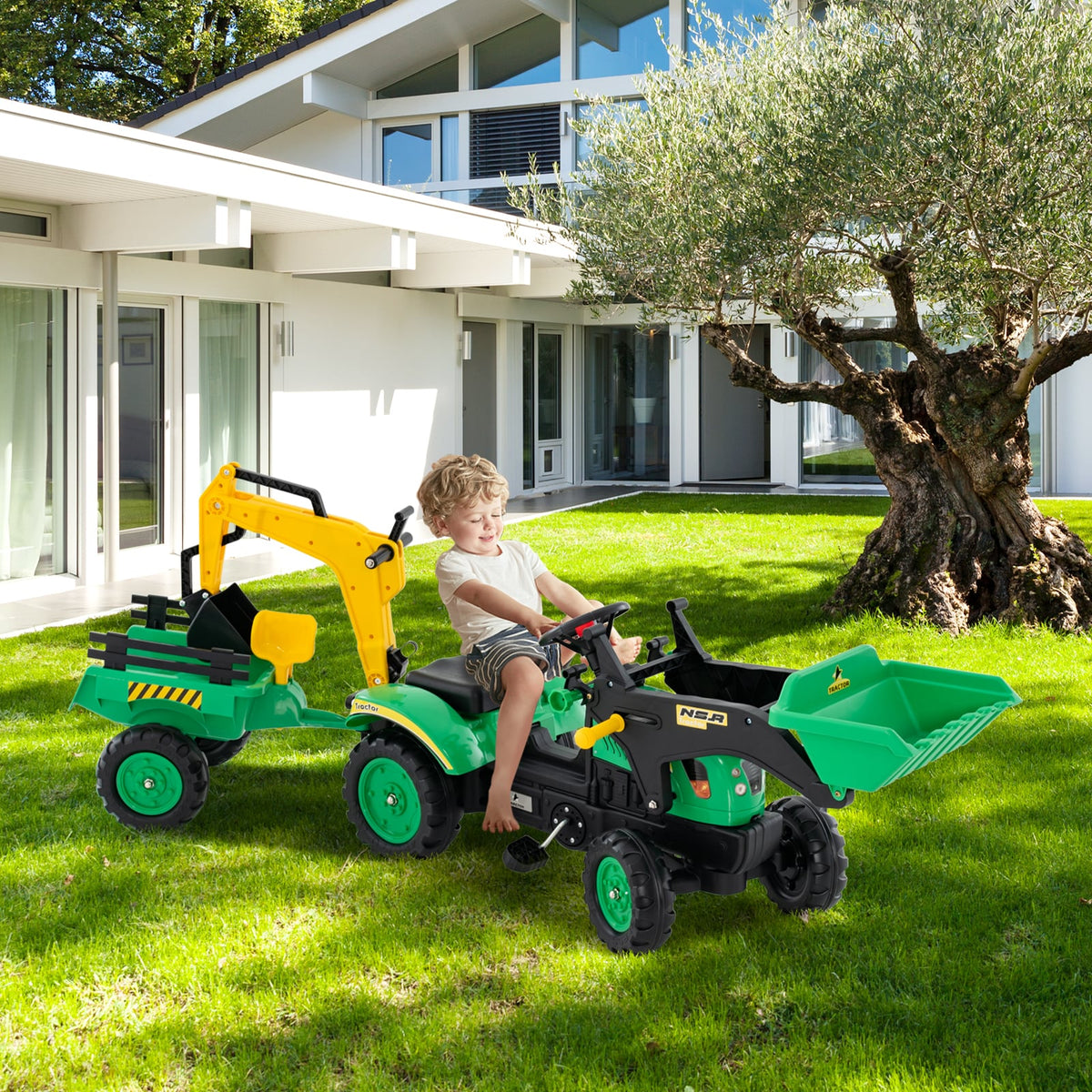 A young boy rides the green pedal excavator with trailer in a garden setting