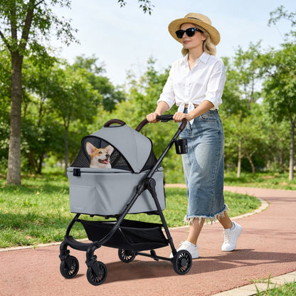 A woman pushes the grey dog stroller with a corgi inside along a path in a park