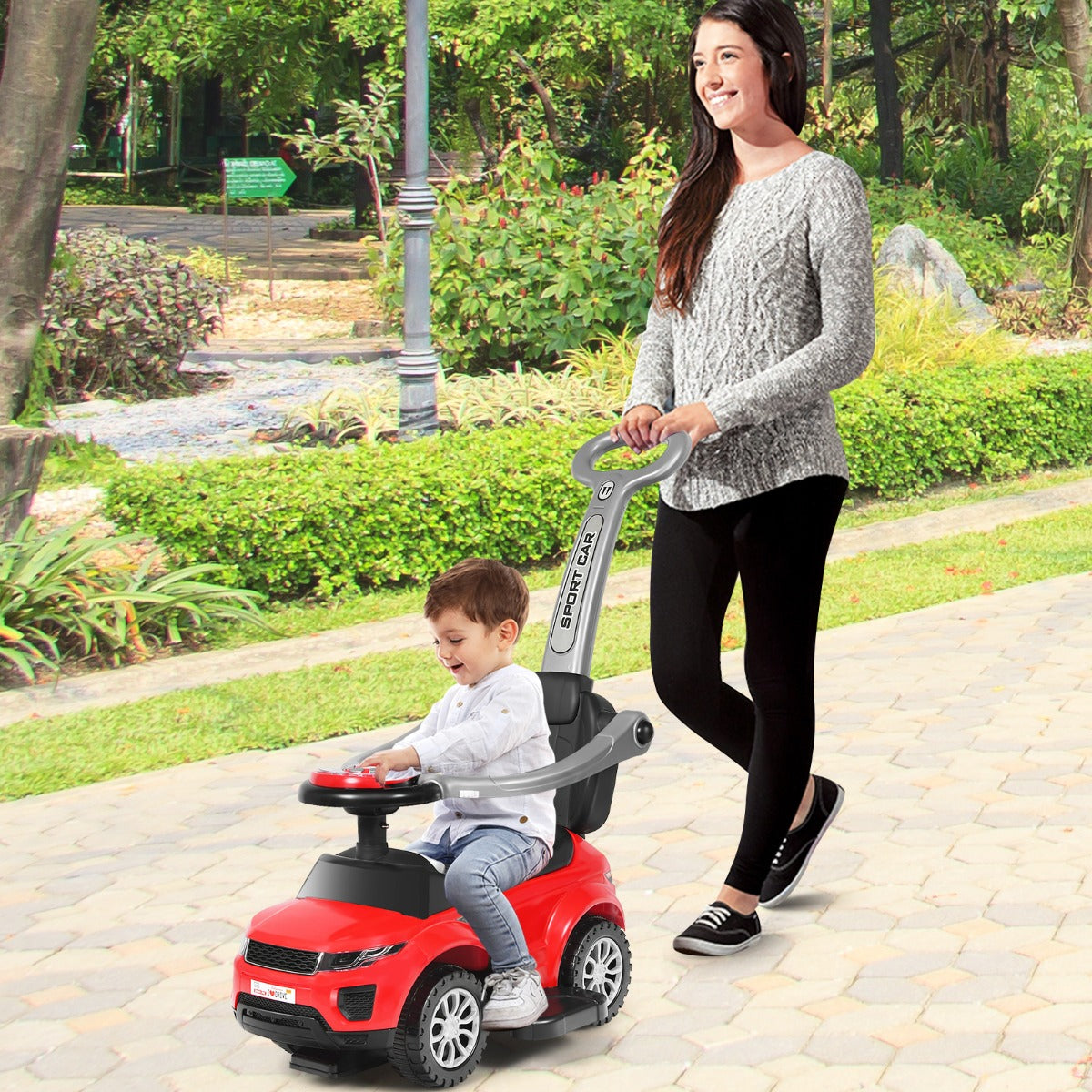 Mother pushing her child on a red ride on push car stroller in a park