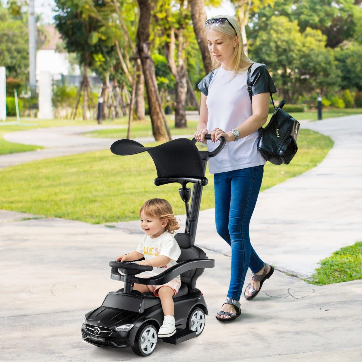A woman pushes a toddler in the black ride on push car in a park