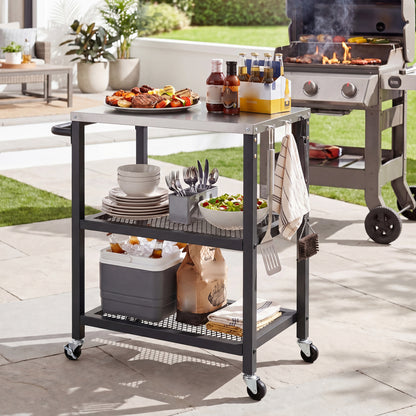The rolling grill cart with a stainless steel top, used for an outdoor barbecue