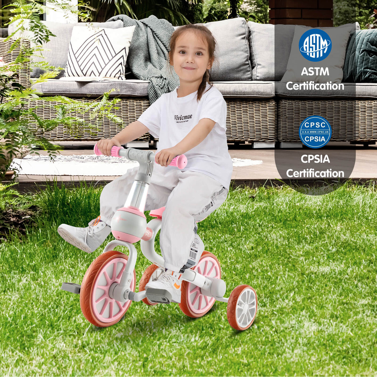 A young girl rides the pink training trike outdoors on grass with cushions in the background