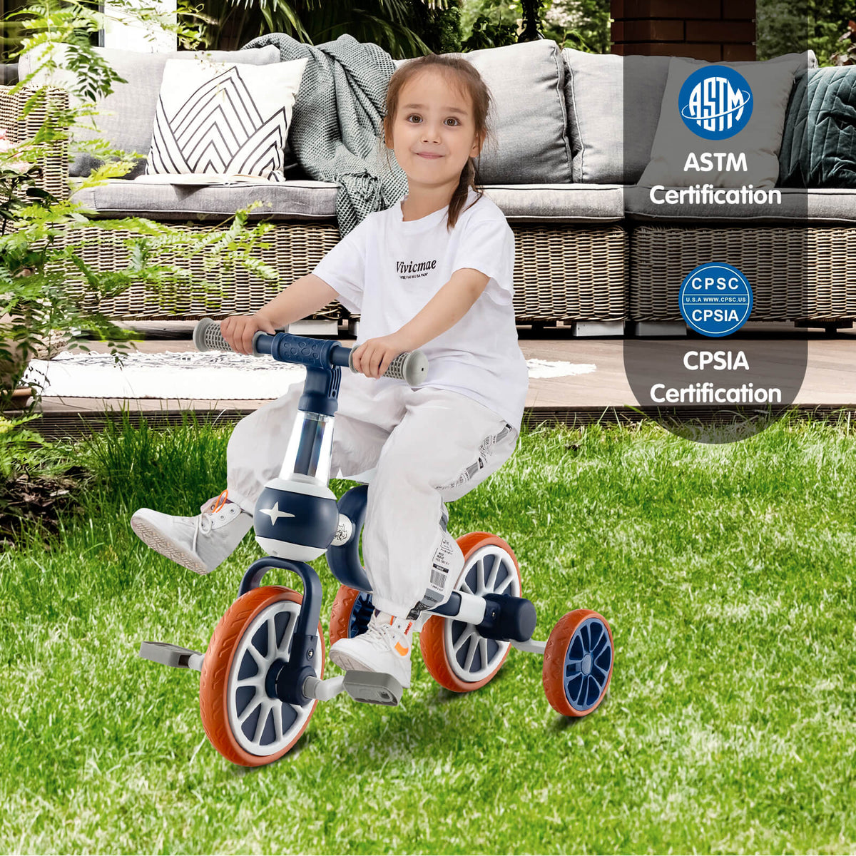 A young girl happily rides the navy training trike on a grassy lawn with outdoor furniture