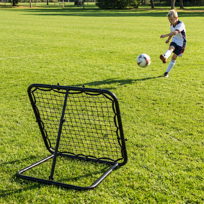 A young boy practices kicking a football against the bounce back net on a grassy field