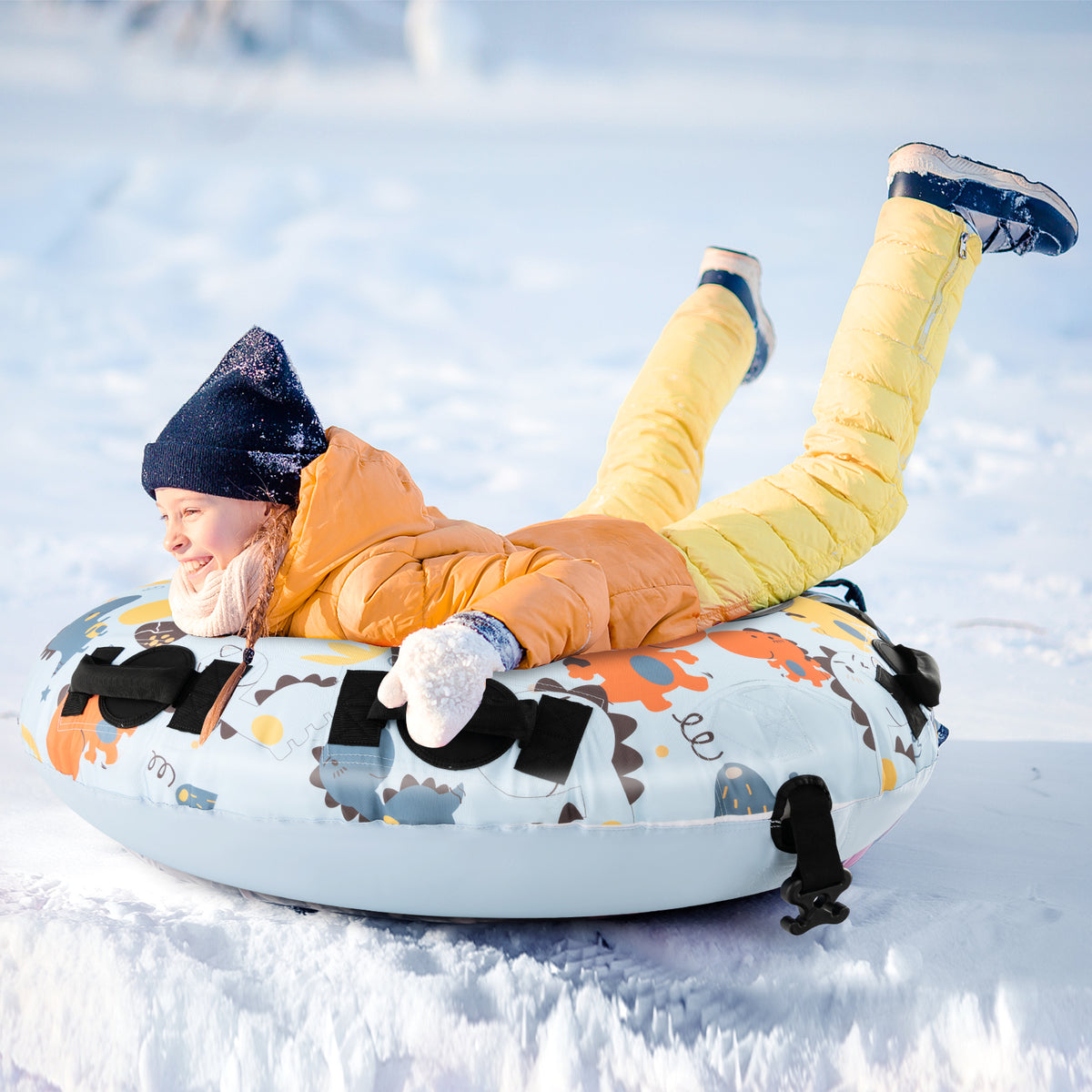 A child wearing yellow snow pants and a yellow jacket lying on the blue dinosaur patterned snow tube in the snow