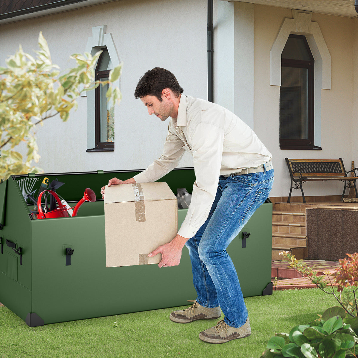 Man placing a cardboard box into the green outdoor storage box, which is open and sitting on grass outside a house