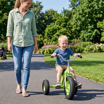 A woman walks beside a young boy on a green kids tricycle in a park