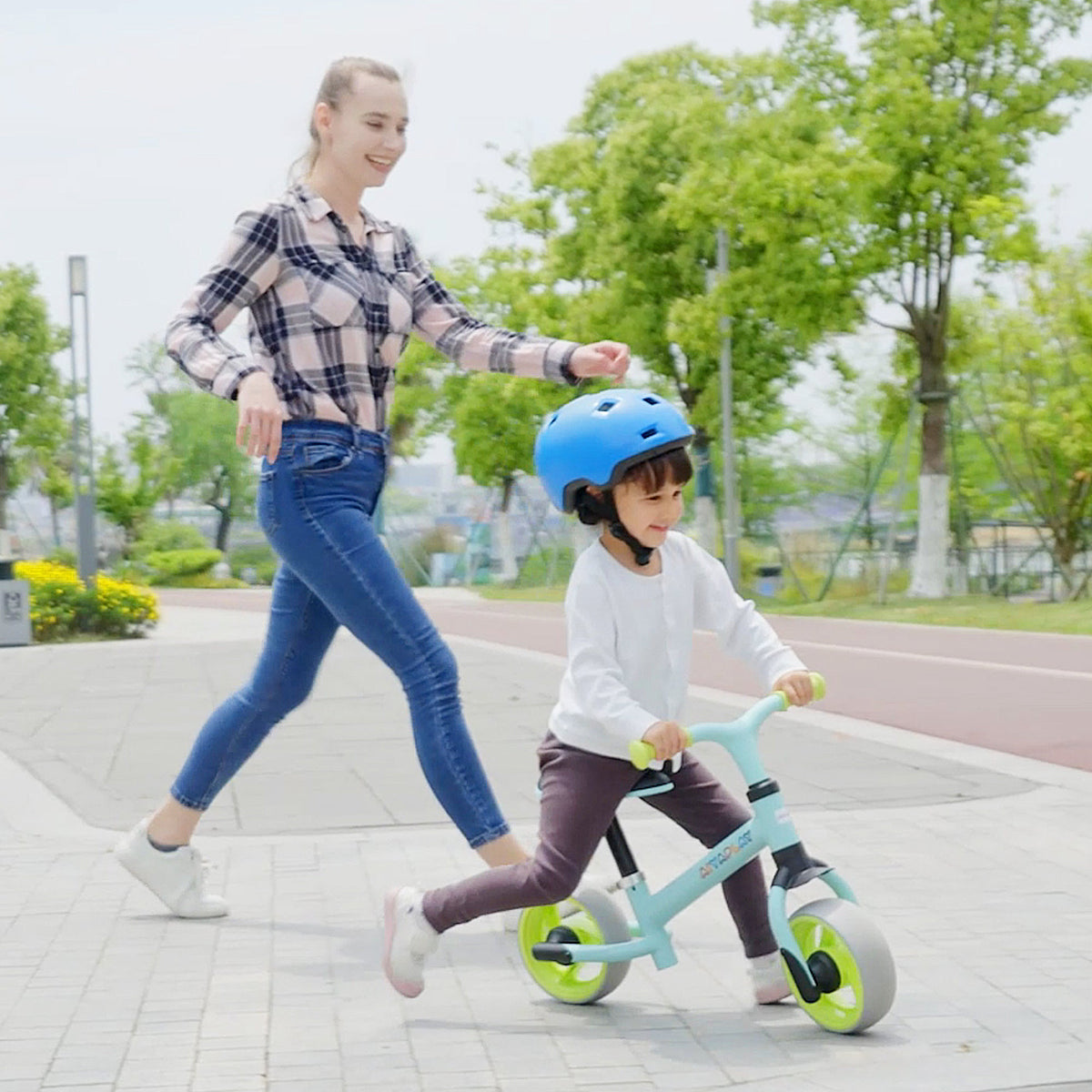 A woman pushes a child on the green balance bike as they ride along a path outdoors