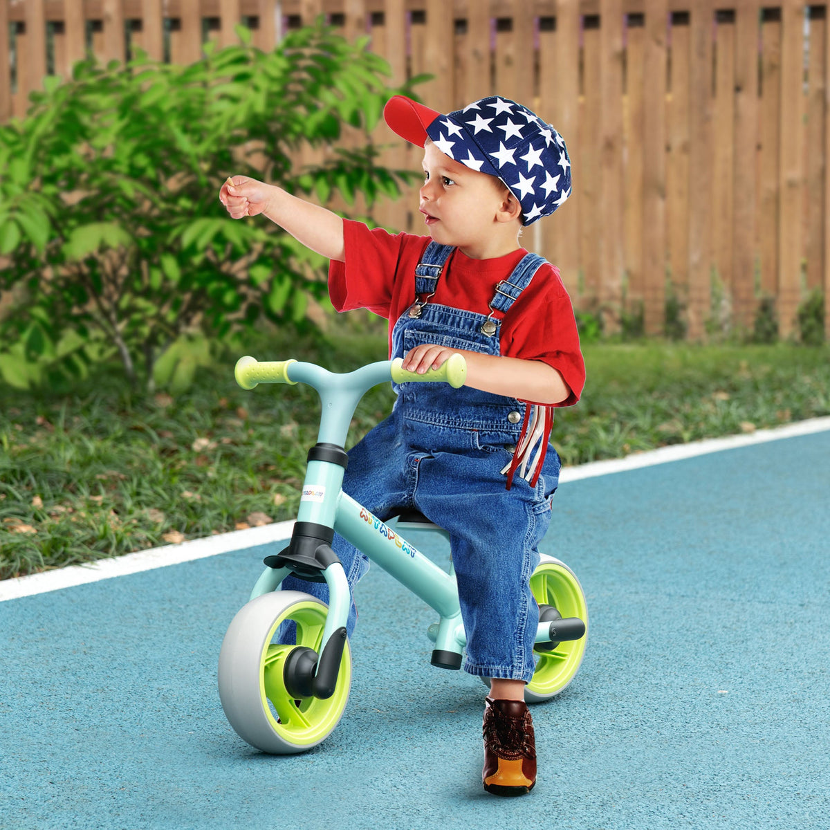 A young child wearing a star-patterned hat rides the green balance bike outdoors on a paved surface