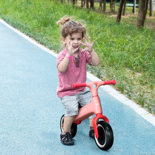 A young child happily riding the pink baby balance bike on a paved path outdoors