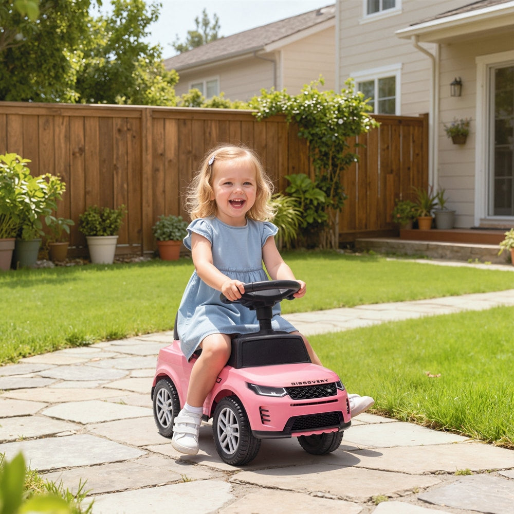 A happy toddler girl rides the pink Land Rover car on a paved garden path
